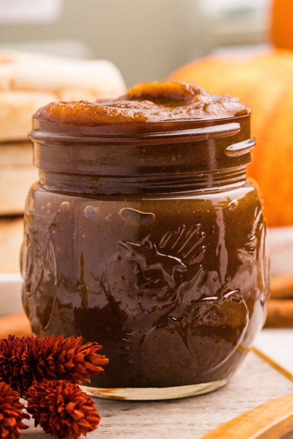 Overflowing jar of glossy dark pumpkin butter beside dried rust-colored flowers with a pumpkin blurred behind.