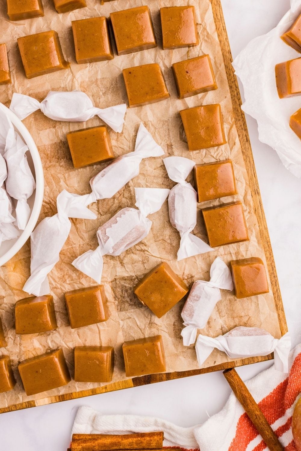 Golden apple cider caramel squares and twist-wrapped pieces on parchment-lined wooden board with cinnamon sticks nearby.